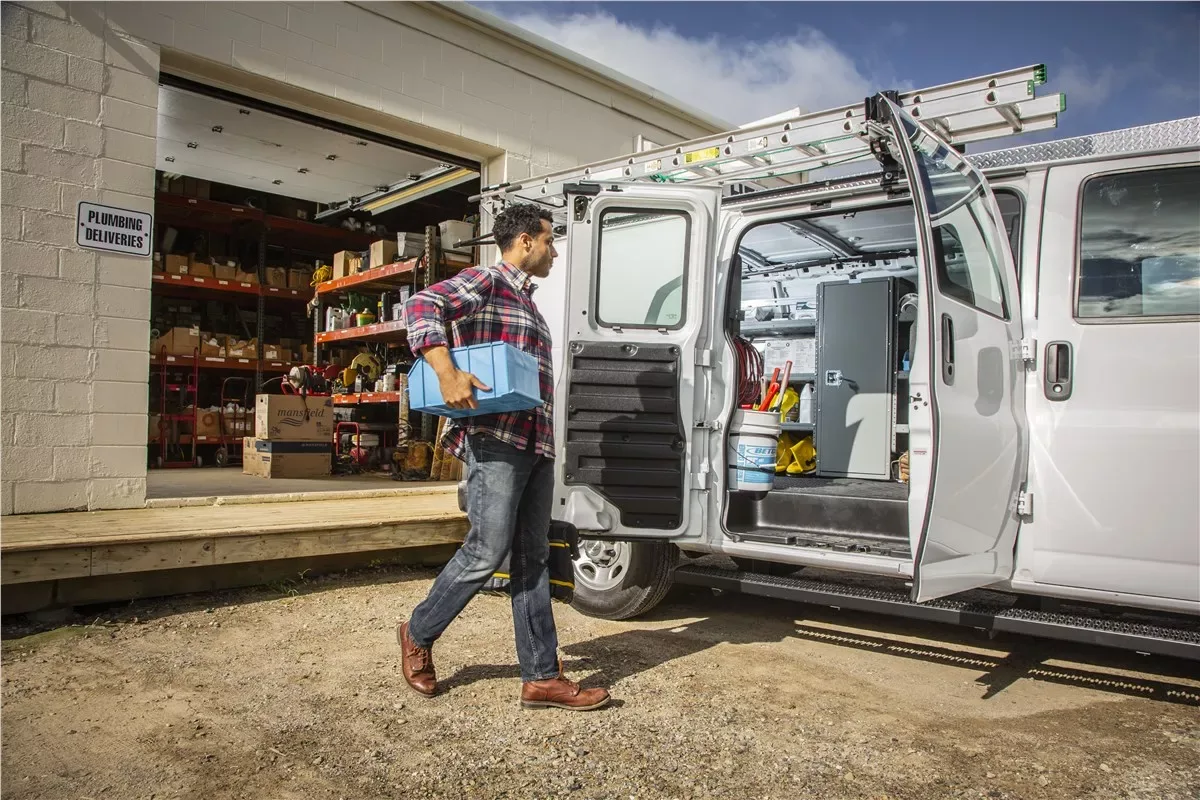 A plumber carrying a parts bin to a shelved cargo van with a roof ladder rack outside a supply warehouse