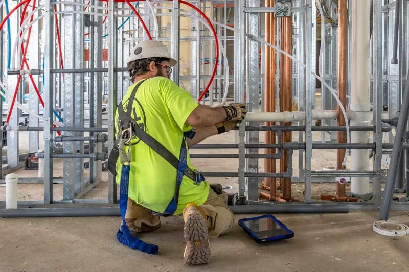 A plumber in a hard hat and safety harness fitting copper and PVC supply lines into metal-stud framing at a commercial site