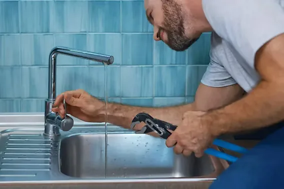 A plumber using a wrench to fit a kitchen faucet over a stainless-steel sink