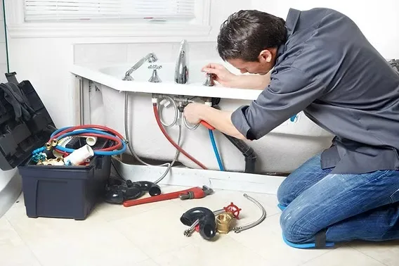 A plumber kneeling to tighten the supply connections under a bathroom sink beside an open tote of wrenches and fittings