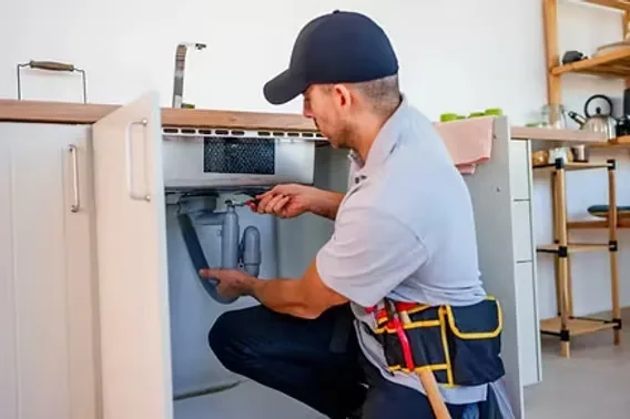 A plumber in a tool belt kneeling to service the drain and supply lines under a modern kitchen sink