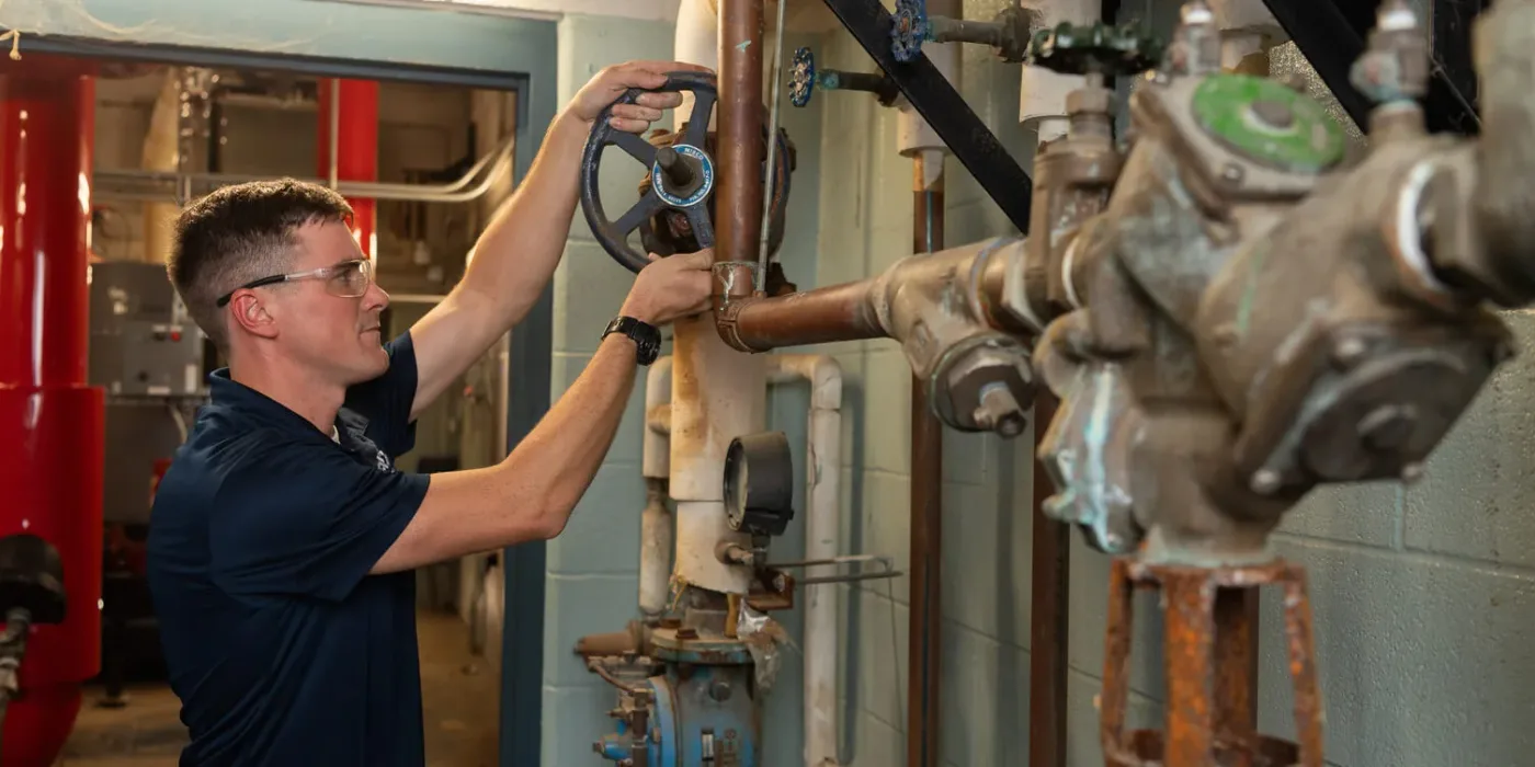 A plumber in safety glasses turning a gate valve on copper riser piping in a commercial mechanical room