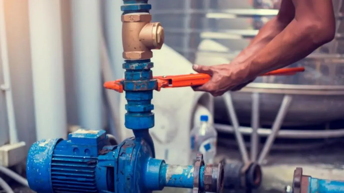 A plumber using a pipe wrench on a bronze valve above an industrial water pump — plumbing contractor insurance in Oklahoma