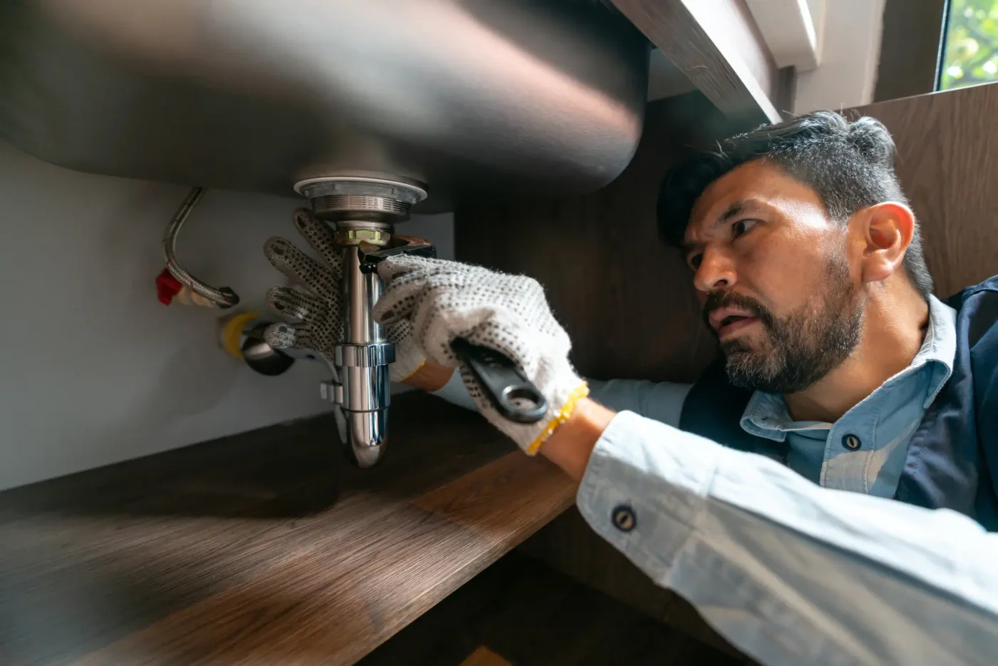 A plumber in work gloves loosening the chrome drain trap under a sink — plumbing contractor insurance in New Mexico