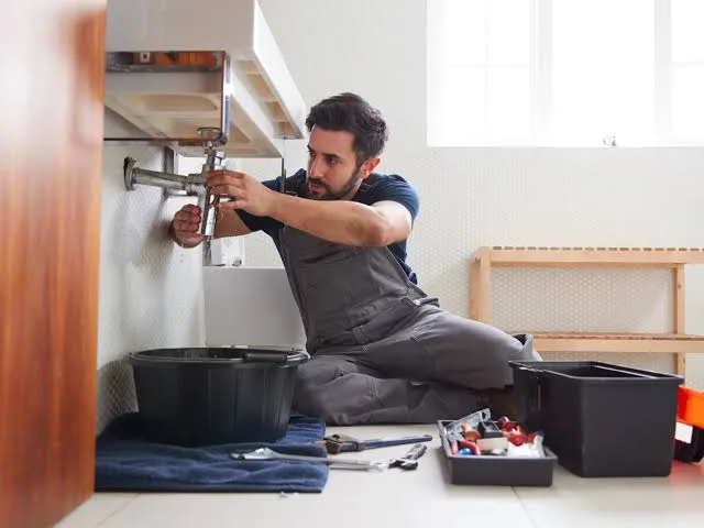 A plumber kneeling to service the trap under a wall-hung sink beside catch buckets — plumbing contractor insurance in Oregon