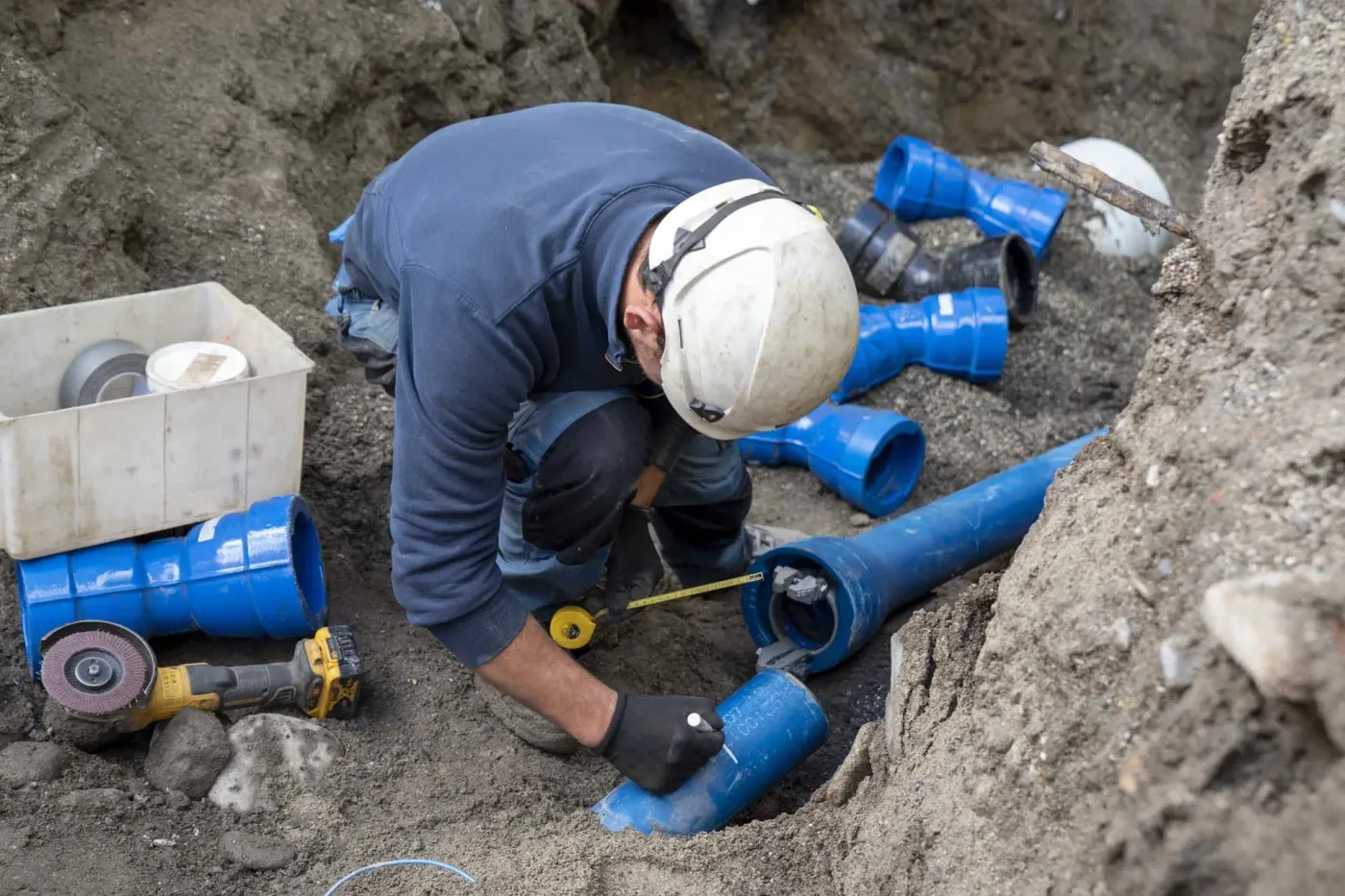 A plumber measuring and marking blue PVC water-main pipe in an open excavation — plumbing contractor insurance in Idaho