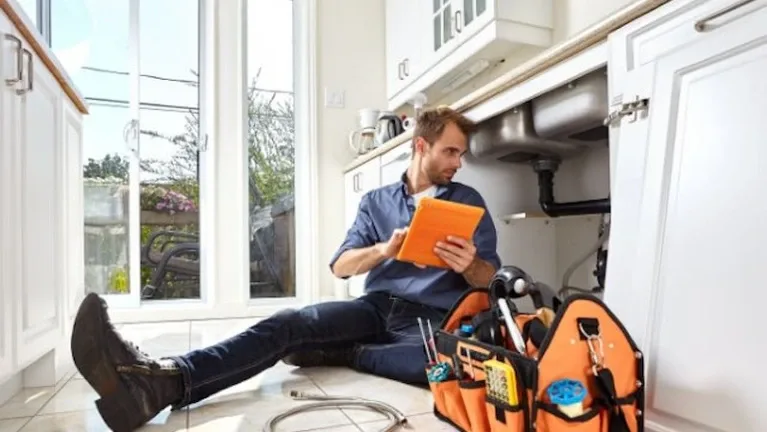 A plumber seated on a kitchen floor reviewing a tablet beside an open tool tote — plumbing contractor insurance in Washington