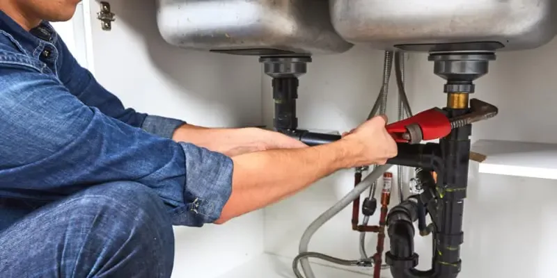 A plumber tightening the drain connections under a stainless double-bowl kitchen sink — plumbing contractor insurance in Michigan